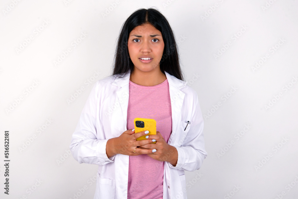 Photo of astonished crazy Young hispanic doctor girl wearing coat over white background hold smartphone dislike feedback concept Photo of astonished crazy Young hispanic doctor girl wearing coat over white background hold smartphone dislike feedback concept
