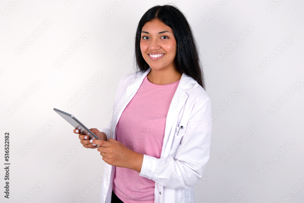 Photo of optimistic Young hispanic doctor girl wearing coat over white background hold tablet Photo of optimistic Young hispanic doctor girl wearing coat over white background hold tablet