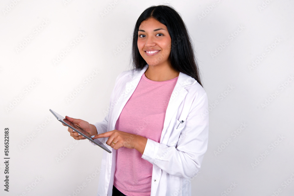 Photo of happy cheerful smart Young hispanic doctor girl wearing coat over white background hold tablet browsing internet Photo of happy cheerful smart Young hispanic doctor girl wearing coat over white background hold tablet browsing internet