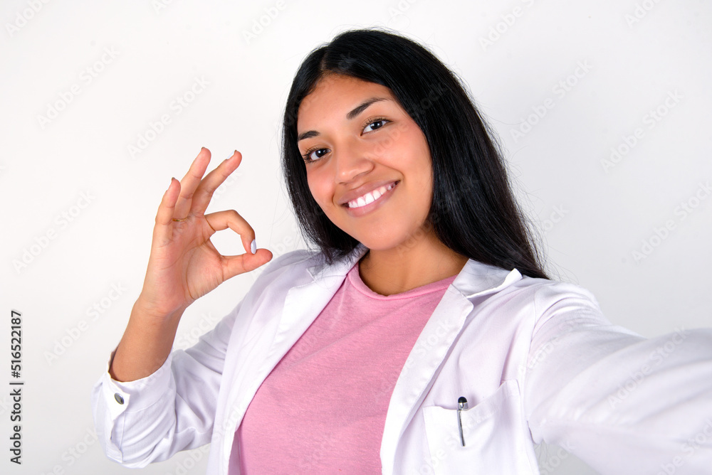 Portrait of pretty cheerful Young hispanic doctor girl wearing coat over white background make selfie show okey symbol Portrait of pretty cheerful Young hispanic doctor girl wearing coat over white background make selfie show okey symbol