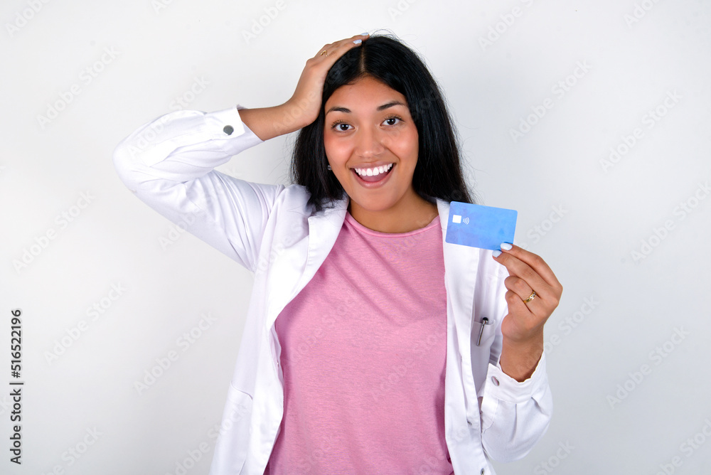 Impressed funny Young hispanic doctor girl wearing coat over white background hand on head holding bank card Impressed funny Young hispanic doctor girl wearing coat over white background hand on head holding bank card