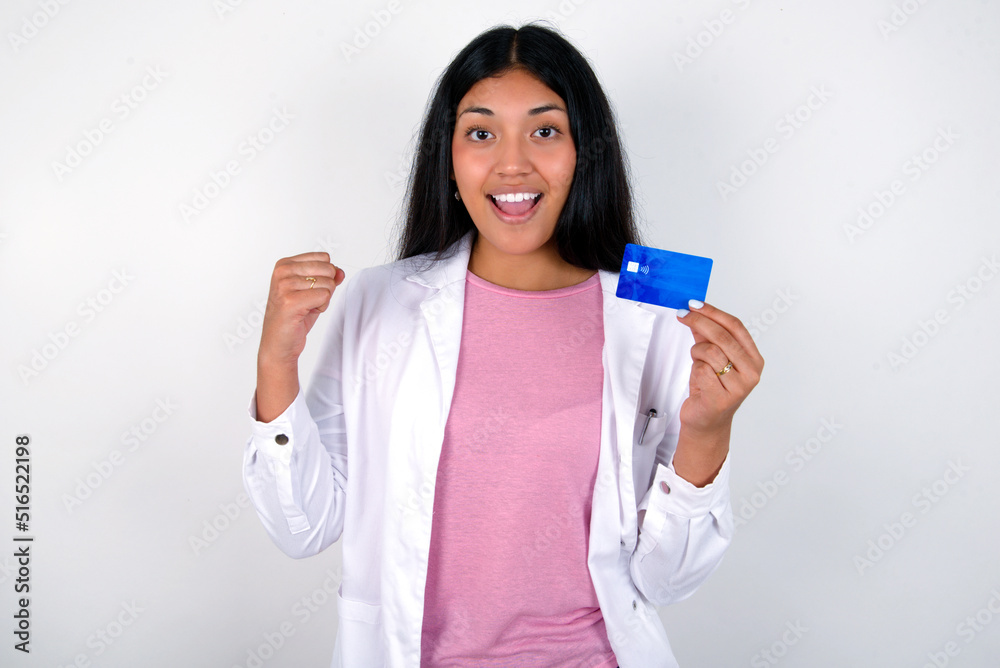 Photo of lucky impressed Young hispanic doctor girl wearing coat over white background arm fist holding credit card. Celebrated Photo of lucky impressed Young hispanic doctor girl wearing coat over white background arm fist holding credit card. Celebrated