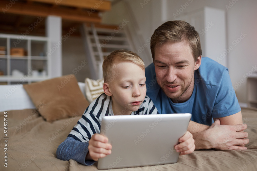 Serious blond boy lying on bed and using digital tablet while watching online movie with father Serious blond boy lying on bed and using digital tablet while watching online movie with father