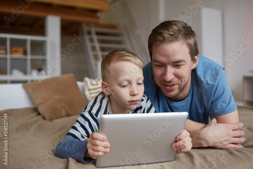 Serious blond boy lying on bed and using digital tablet while watching online movie with father