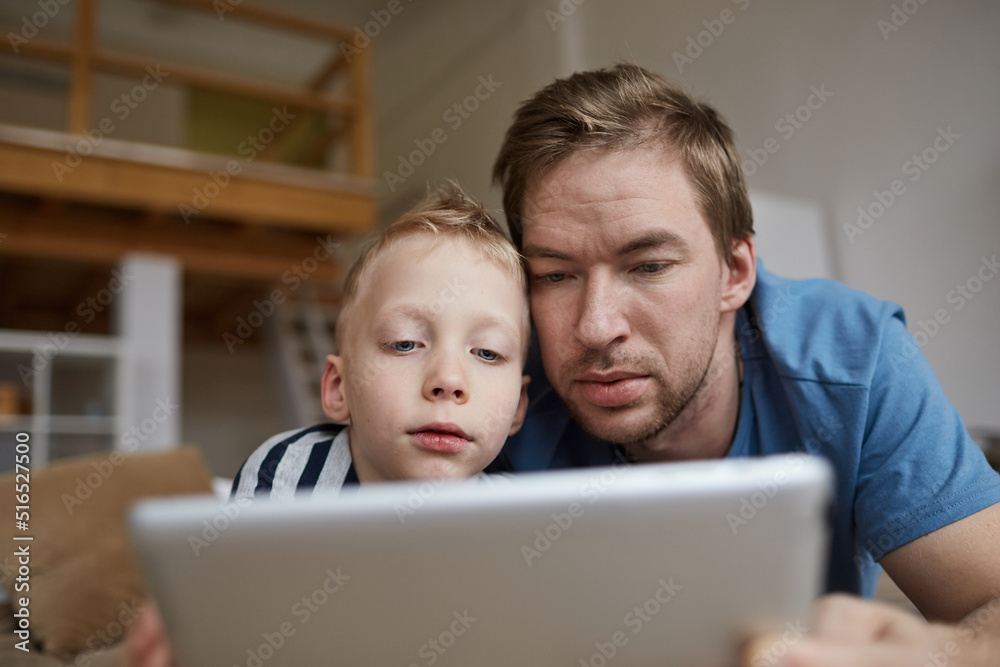 Close-up of serious curious young father and son lying on bed at home and watching film on digital tablet