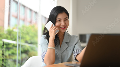 Confident Asian businesswoman having phone conversation with business partners and using laptop at modern office