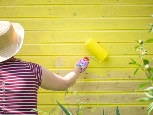 young girl paints a wooden wall with a roller with yellow paint