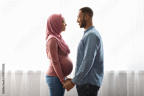 Married pregnant muslim couple standing near window at home and holding hands