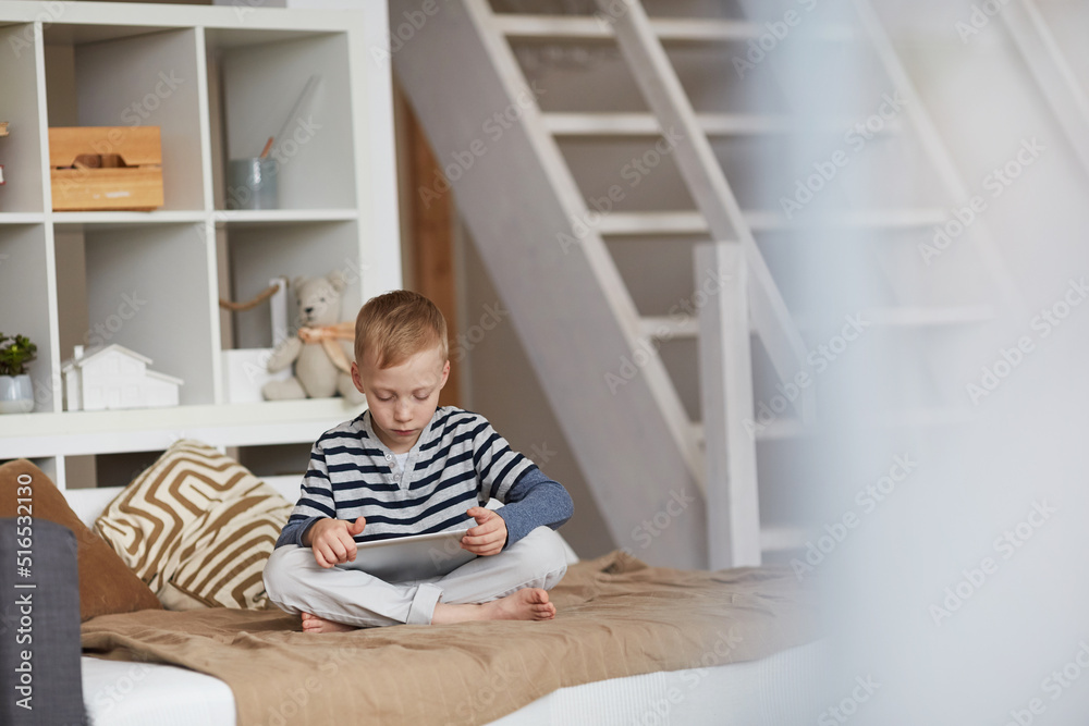 Serious little boy with blond hair sitting with crossed legs on bed and performing online tasks on tablet Serious little boy with blond hair sitting with crossed legs on bed and performing online tasks on tablet