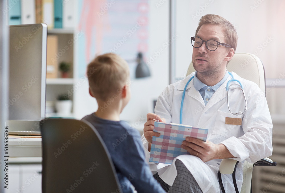 Content handsome young pediatrician with stethoscope around neck making notes in medical file while asking little boy about health complaints