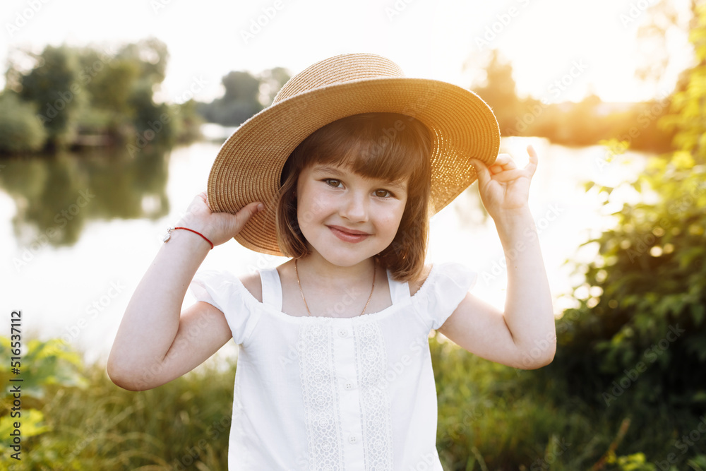 Little smiling girl in straw hat is having fun on summer holiday vacations. Happy child playing and looking in camera by the lake. Kid having fun outdoors. healthy lifestyle concept.