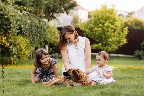 Family day, mother's day. Beautiful smiling young mom and two child daughters cuddling happy domestic dog on the backyard lawn. Idyllic family having fun with pet outdoors on summer holiday.