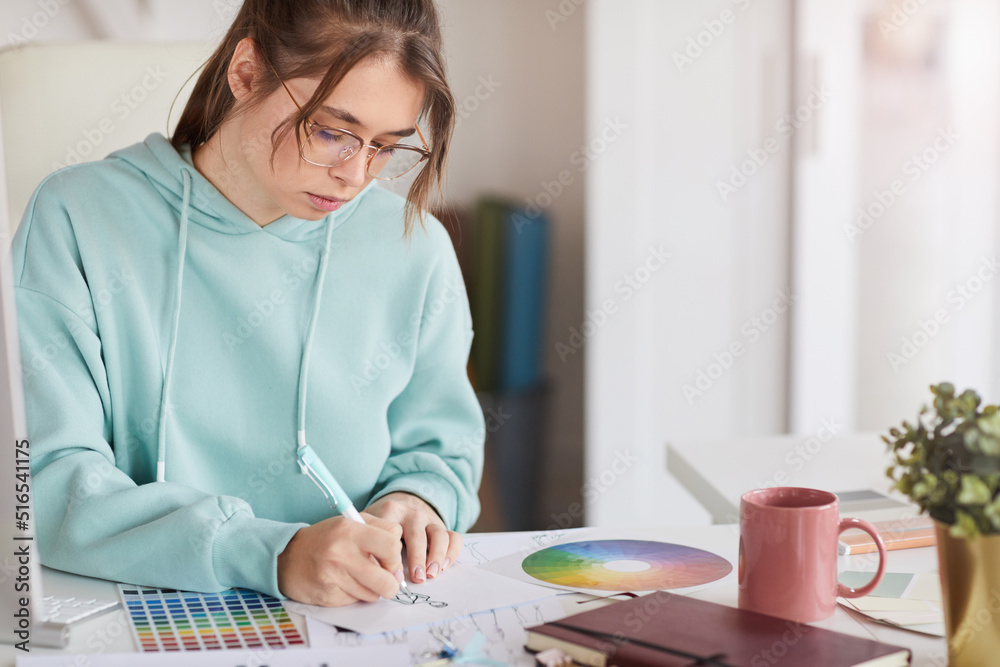 Serious concentrated young female designer in teal hoodie sitting at table and drawing fashion sketch with pen Serious concentrated young female designer in teal hoodie sitting at table and drawing fashion sketch with pen