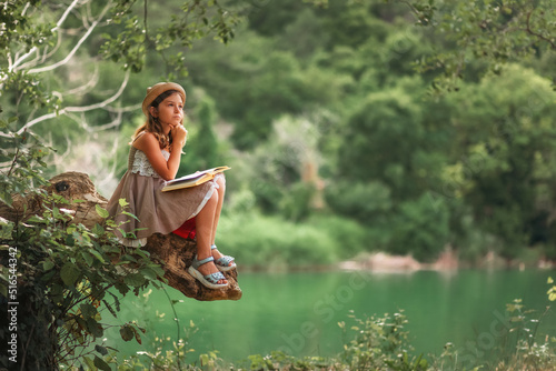 Pensive schoolgirl in a dress and a straw hat reading a book sitting on a old log. In the background are park and river. Copy space. The concept of children's education and school holidays