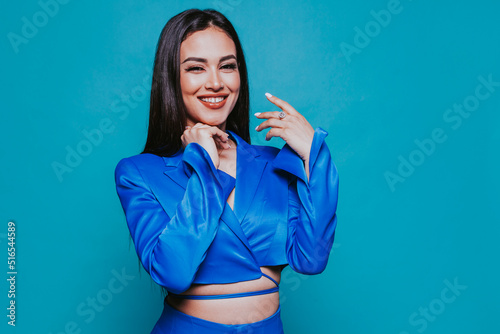 Young happy smiling arabic girl wears blue suit  poses with raised hand shows ring  over blue background at studio. Fashion  stylish attractive woman.