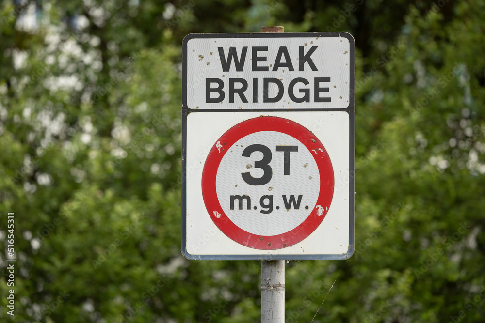 Outdoor white sign on a black pole showing a weak bridge with symbols and numbers