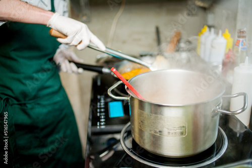 The cook in an apron and gloves is stirring the soup in a saucepan. Chef cooking a delicious dish in the kitchen. The owner of the soup kitchen or field kitchen interferes with the bucket in the soup.