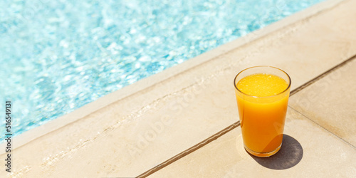 Glass of fresh cool orange juice drink on swimming pool board, summer tropical holiday concept.Banner with copy space