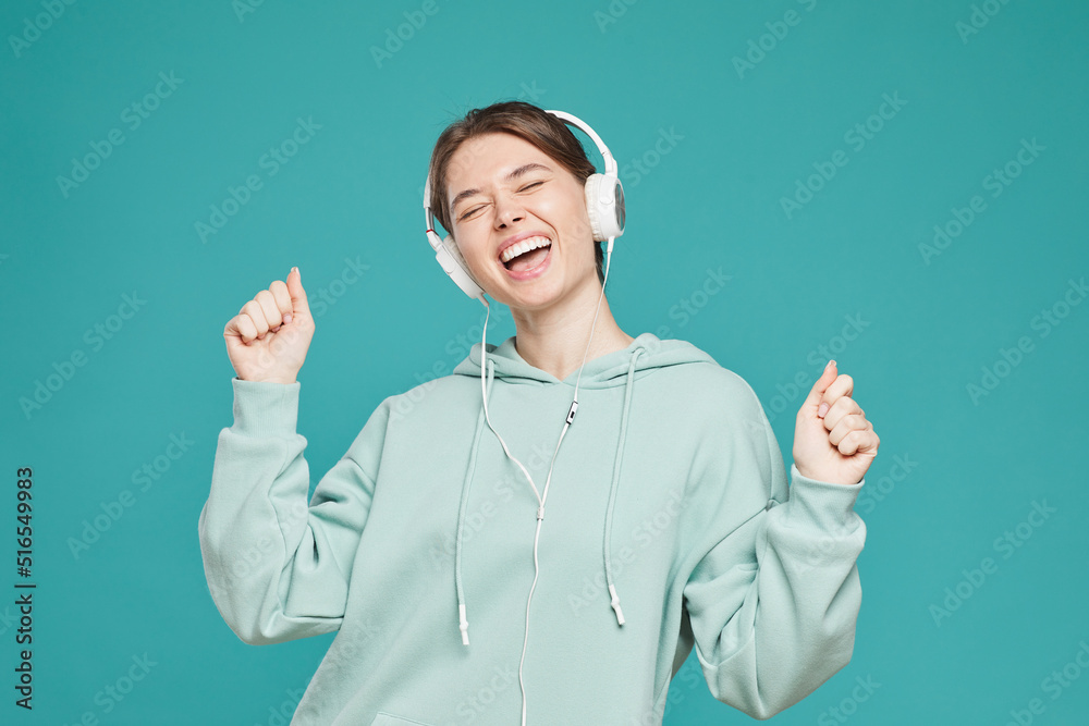 Ecstatic pretty girl in headphones and hipster hoodie standing against teal background and dancing with closed eyes