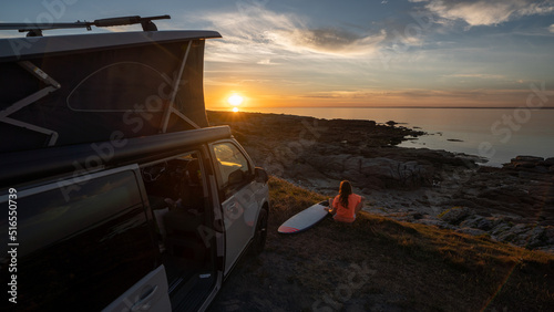 Photographie Surfer girl sitting near her mini van and looking on the ocean at summer sunset