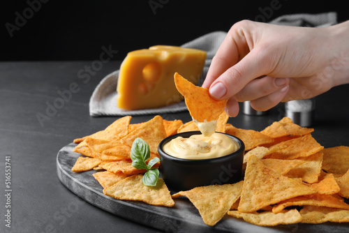 Fotografie Woman dipping crispy nacho into delicious cheese sauce at black table, closeup