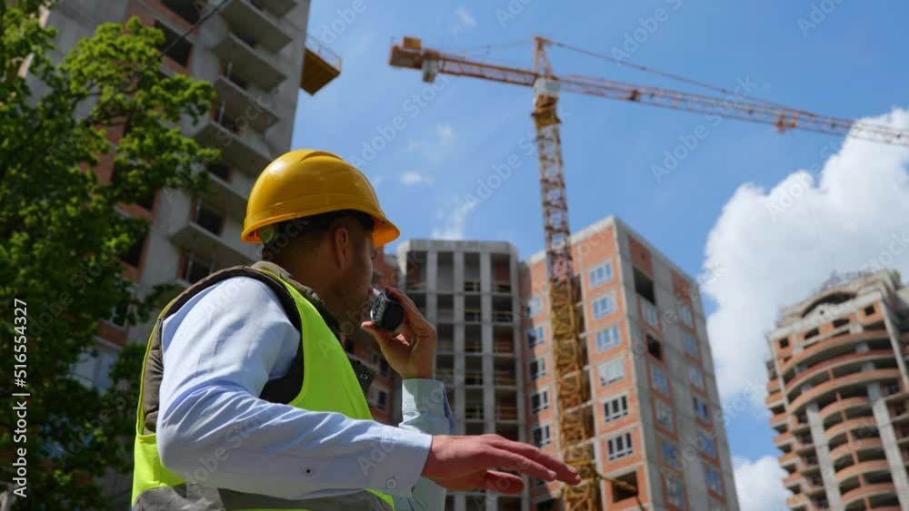 Close up of handsome male worker in hard hat talking in walkie-talkie standing at construction site on street outdoor. Low angle view of man constructor engineer using receiver at work Close up of handsome male worker in hard hat talking in walkie-talkie standing at construction site on street outdoor. Low angle view of man constructor engineer using receiver at work