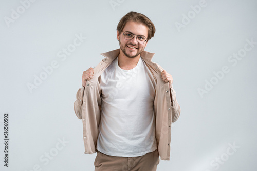 Handsome charismatic guy with glasses. Smiling cheerful man stands on gray background and holds edges of open jacket with hands, showing t-shirt. Place for design, logo, inscription, advertisement © VSH-PRODUCTION