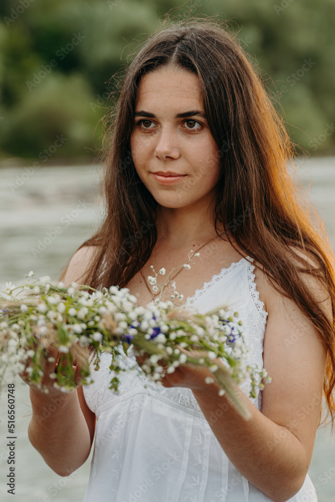 Caucasian girl in long white dress stands in shallow river and holds wreath of wild flowers in hands. River, forest and cloudy sky at sunset.Traditional rite of fortune-telling on feast of Ivan Kupala