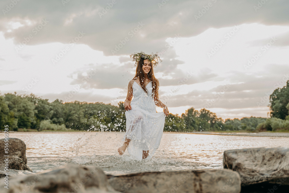 Cheerful girl at sunset is standing in shallow river in long white dress and with wreath on head and having fun. Woman splashes water with foot and laughs. On holiday of Ivan Kupala girls are guessing Cheerful girl at sunset is standing in shallow river in long white dress and with wreath on head and having fun. Woman splashes water with foot and laughs. On holiday of Ivan Kupala girls are guessing