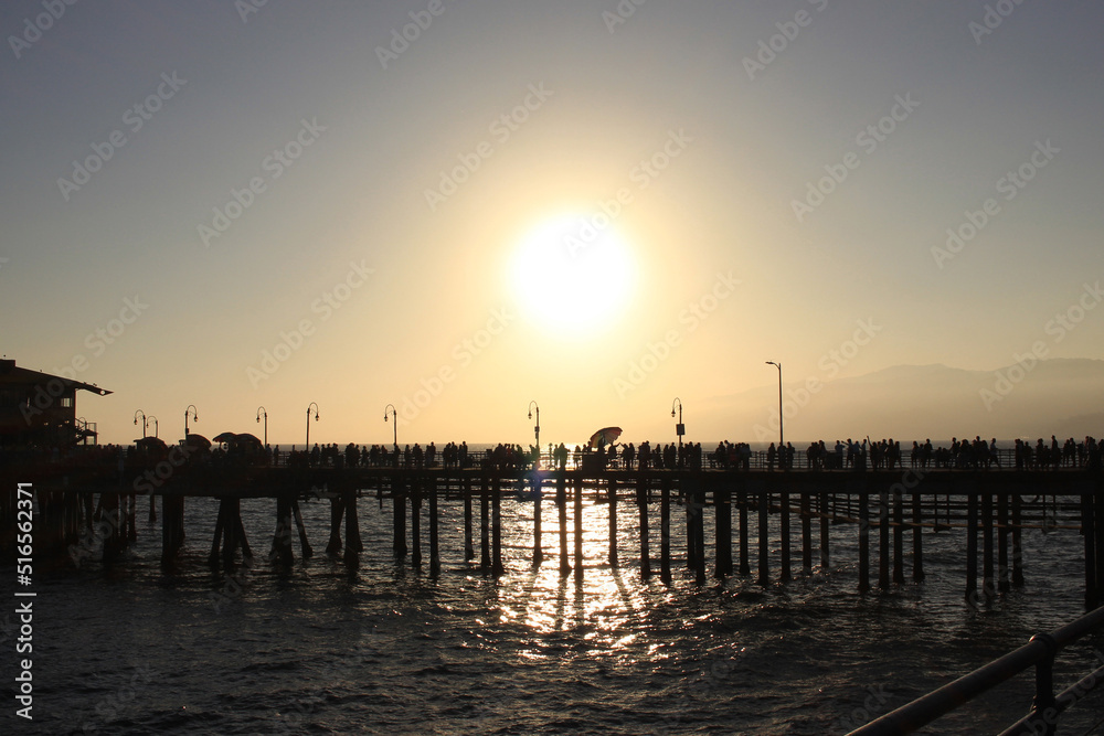 Backlit silhouettes of many unrecognizable people walking across the Santa Monica Pier at a slightly misty sunset with the sparkling sea