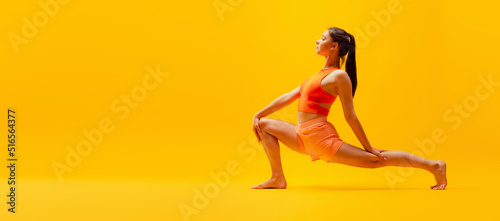 Portrait of young slim, flexible girl in shorts doing stretching exercises isolated on bright yellow background. Sport, health, active lifestyle