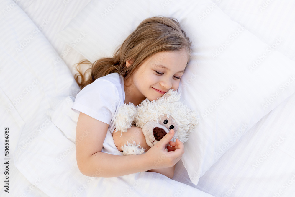 cute baby girl sleeping sweetly on the bed on a white cotton pillow under the blanket and smiling, hugging a teddy bear toy, healthy baby sleep at night cute baby girl sleeping sweetly on the bed on a white cotton pillow under the blanket and smiling, hugging a teddy bear toy, healthy baby sleep at night