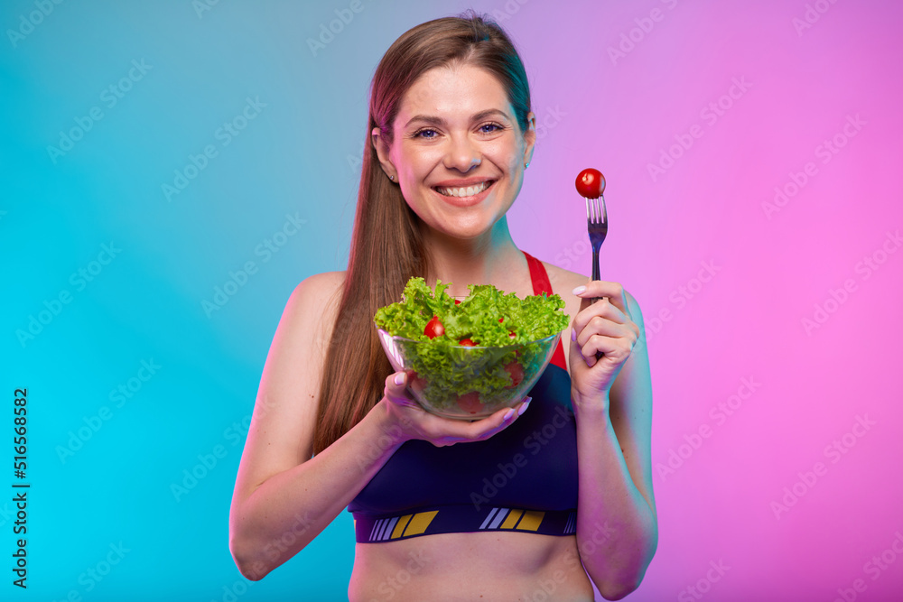 Sporty woman in sportswear green salad in glass bowl and red tomato on fork. Female fitness portrait isolated on neon multicolor background. Sporty woman in sportswear green salad in glass bowl and red tomato on fork. Female fitness portrait isolated on neon multicolor background.