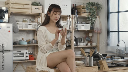 sexy chinese woman sitting on kitchen table with folded leg is checking phone messages in leisure on a tranquil morning at home.