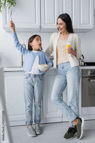 astonished girl holding spoon in raised hand near nanny with glass of orange juice.