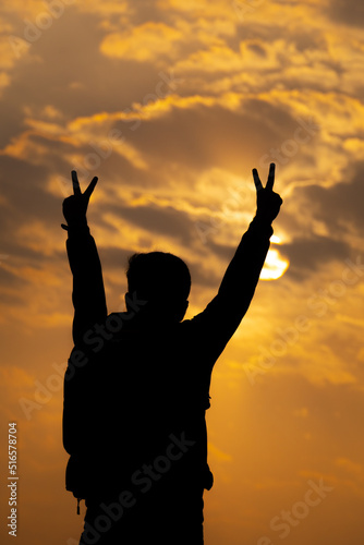Silhouette shot of an Indian hiker wearing backpack doing victory sign with his hands in front of the sun during the sunset. Hiker does V sign after reaching the top of the mountain. Victory concept