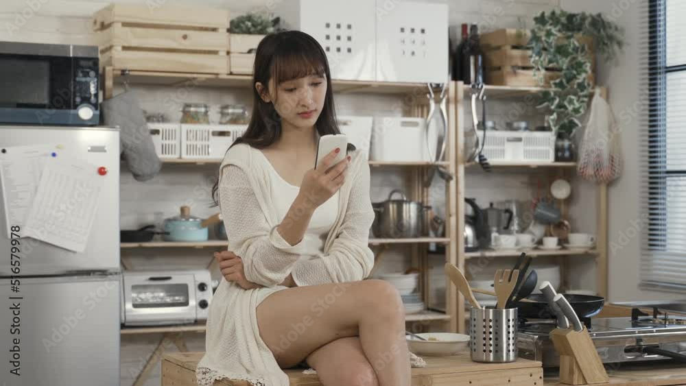 portrait of attractive asian female sitting cross legged on kitchen table is reading online articles on her smartphone with concentration during daytime at home