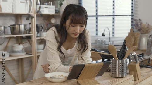 asian girl leaning prone on kitchen table is selecting and watching videos on her touchpad while having a bowl of corn flake with milk for breakfast at home. photo