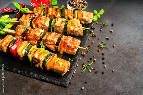 Grilled pieces of chicken meat on skewers.Grilled kebab pieces with vegetables on skewers on a ceramic black board on a dark background with fresh basil leaves.