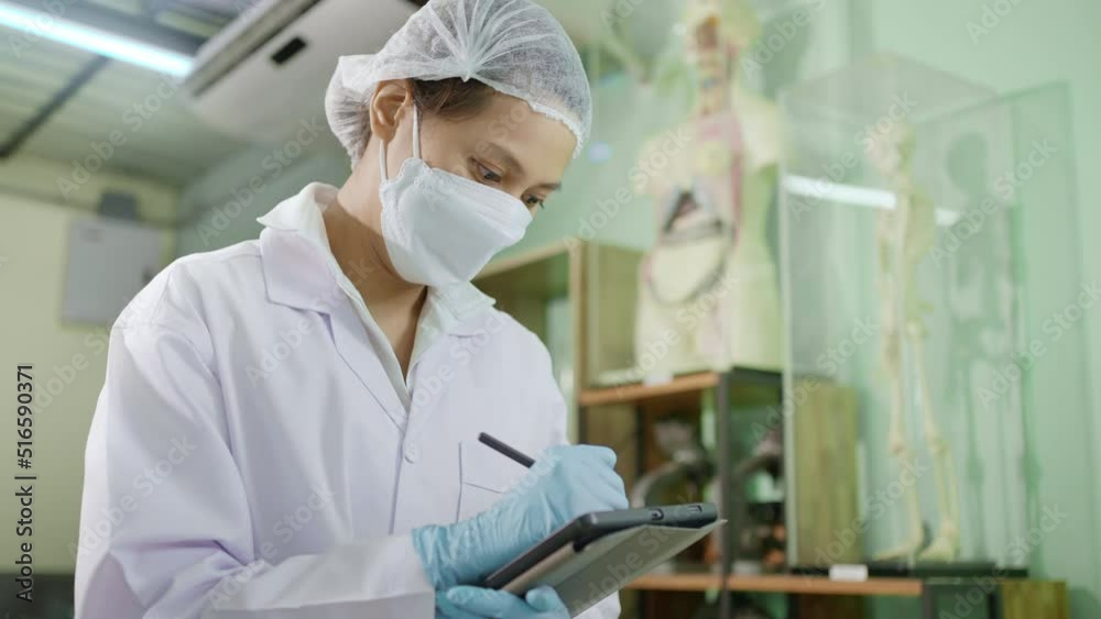 Young female scientist or lab technician in white lab coat wearing safety mask and hair net while working with microscope in her lab and taking notes Young female scientist or lab technician in white lab coat wearing safety mask and hair net while working with microscope in her lab and taking notes