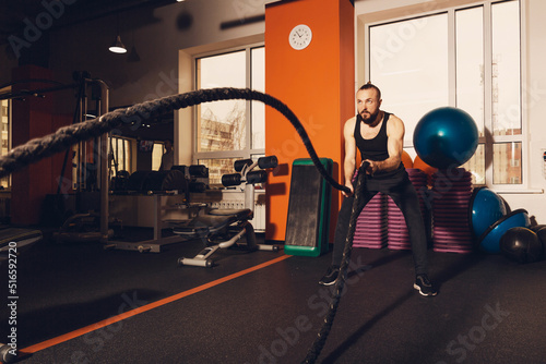 Athletic young man with a beard does exercises with a battle rope does exercises in a functional fitness gym