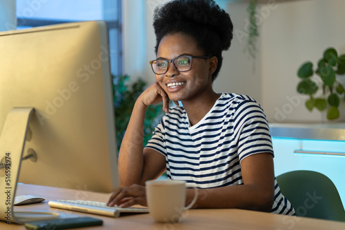 Joyful cheerful African American millennial woman looking at computer monitor, happy young black female sitting at table watching movie and drinking tea, enjoying leisure time in evening at home