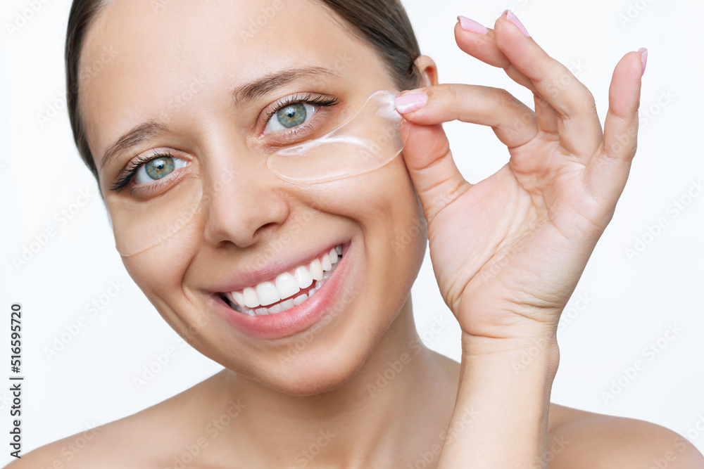 A young attractive caucasian smiling cheerful woman puts gel colorless transparent patches on the skin under her eyes isolated on a white background. Skin care, cosmetology A young attractive caucasian smiling cheerful woman puts gel colorless transparent patches on the skin under her eyes isolated on a white background. Skin care, cosmetology