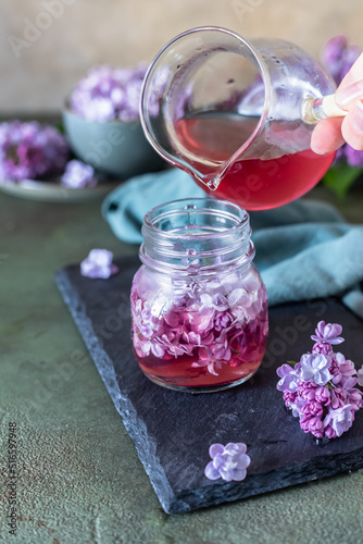 Preparation of syrup from the lilac flowers. Glass jar of homemade lilac syrup and branch of lilac flowers, stone background.