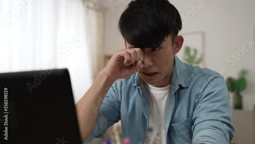 shoulder shot of an overworked asian male employee rubbing his tired eyes while working from home on the laptop in the living room at daytime.