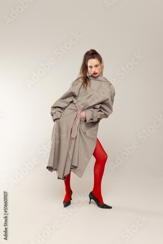Portrait of young stylish girl posing in red tights and coat posing isolated over grey studio background. Blushing cheeks.