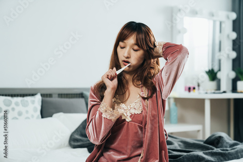 closeup drowsy asian female wearing pajamas is scratching her head with eyes close while brushing her teeth after waking up in a bright home bedroom interior. photo