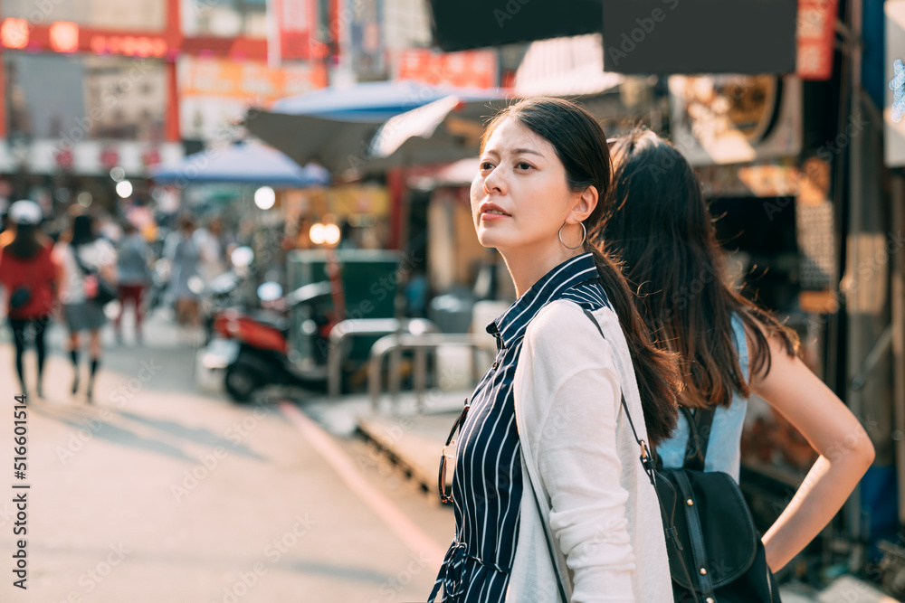 beautiful curious Asian female is turning around to look at the distance while shopping with her friend on the street of a sightseeing business area in the sun.