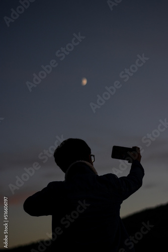 vertical portrait of a young man taking a picture of the Moon with his cell phone at sunset at dusk.