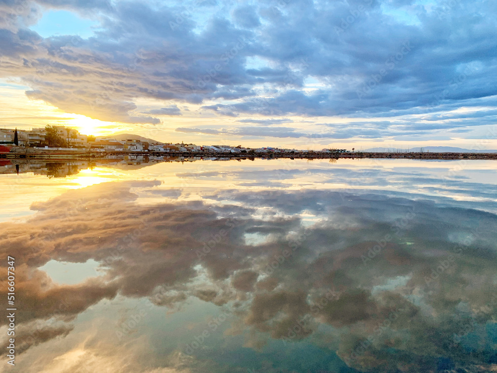 A reflection of the sunset on the sea surface on a small beachside town with clouds in the sky.